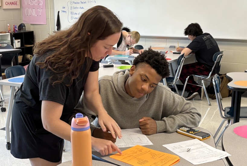 teacher stands next to a seated student and points to their paper